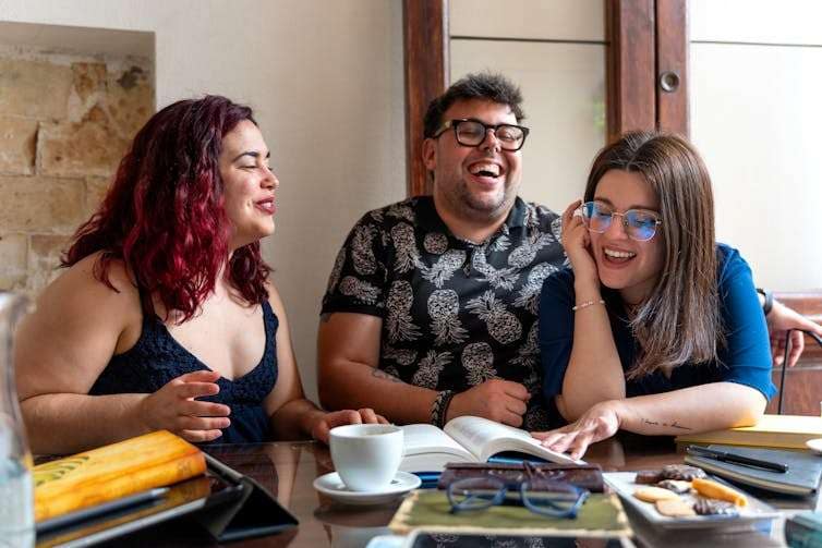 three people laughing together at a table, with books open in front of them