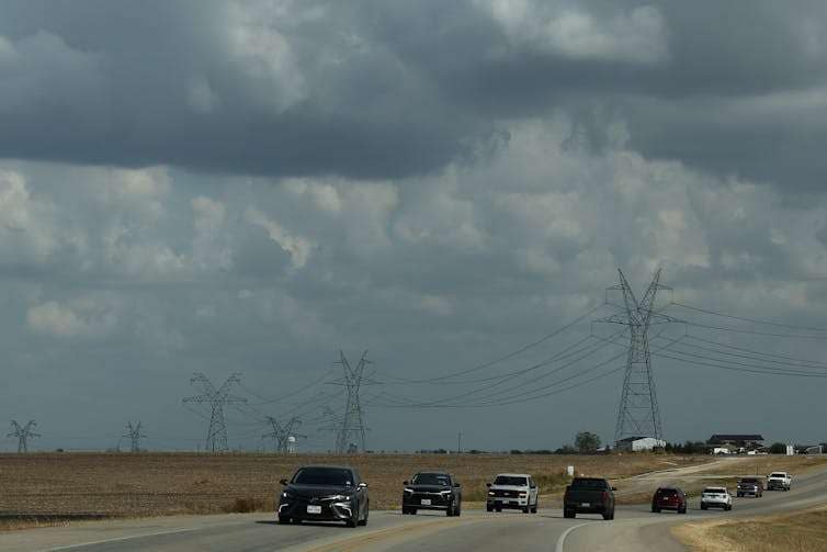 A line of tall metal towers holding wires stretches into the distance over a highway.