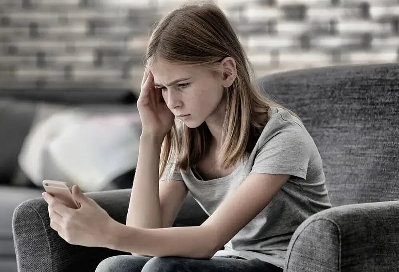 Teenage girl sitting on a couch staring at her phone with a worried expression.